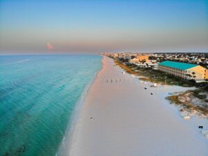 Aerial shot of emerald blue waters along a pink sandy beach. A hotel with a turquoise roof stands long the right with other small buildings.