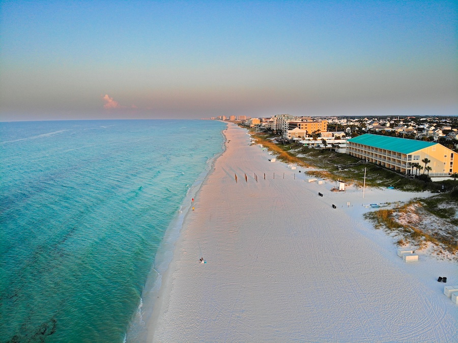 Aerial shot of emerald blue waters along a pink sandy beach. A hotel with a turquoise roof stands long the right with other small buildings. 