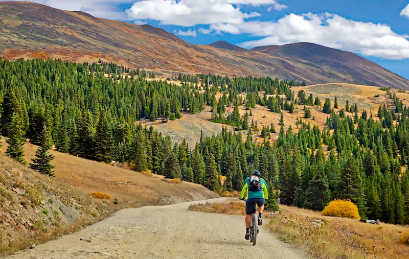 Photo of a cyclist in a green shirt and black backpack riding a bike on a dirt path, with green trees ahead and mountains with blue skies and clouds