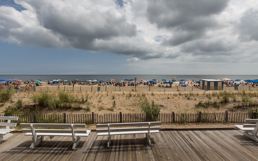 Photo of a boardwalk with cloudy skies looking toward the ocean with people and umbrellas on the beach