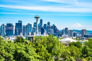 Photo of the skyline of Seattle including the Space Needle on a bright, clear, blue sky day with green trees in the foreground
