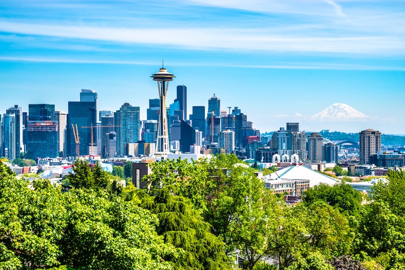 Photo of the skyline of Seattle including the Space Needle on a bright, clear, blue sky day with green trees in the foreground