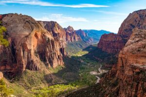 A photo of the Angels Landing Gorge of Zion National Park, with green trees running through surrounded by steep red earth cliffs and a blue sky